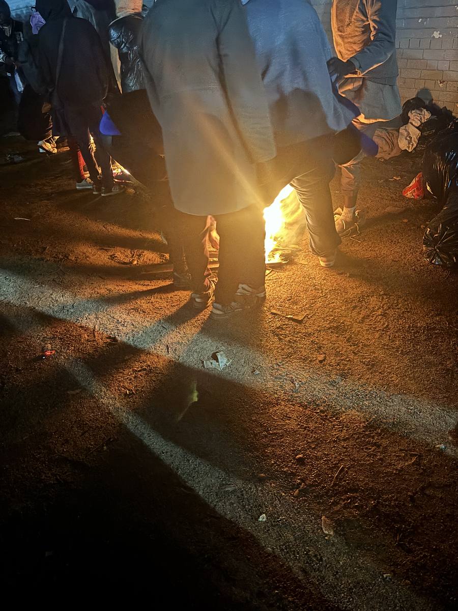 People huddled around a makeshift fire on wet asphalt in the cold night outside Home Affairs